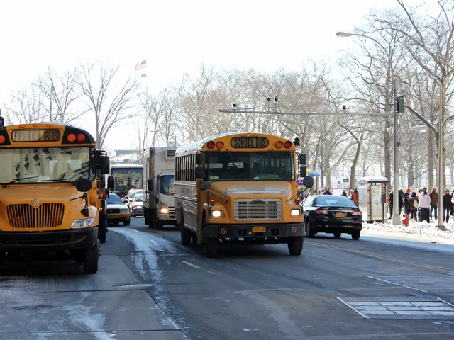 The image shows a group of school buses driving down a street lined with tall buildings. There are...