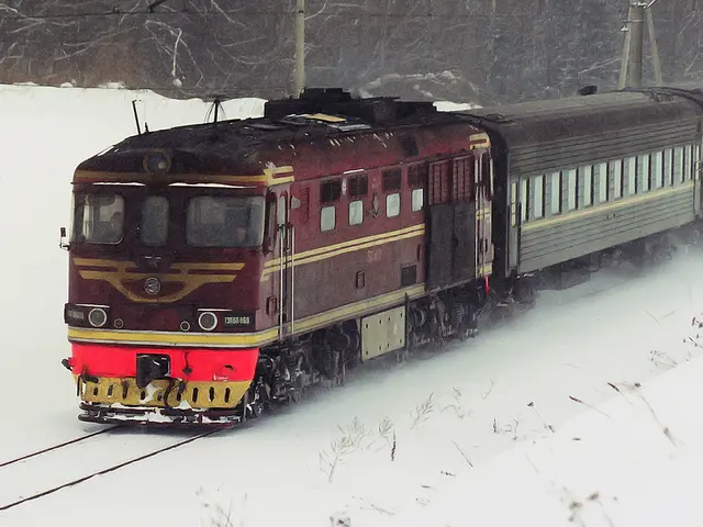 The image shows a train traveling down train tracks covered in snow, with electric poles and trees...
