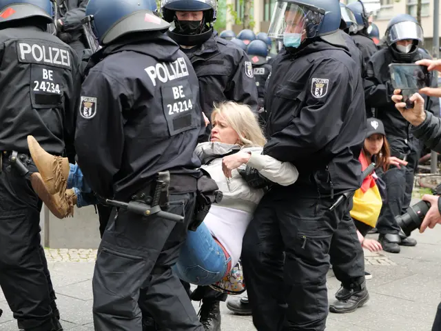 The image shows a group of police officers in riot gear standing around a woman being escorted by...