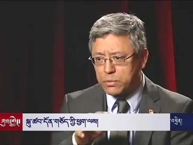 The image shows a man in a suit and tie sitting down, wearing spectacles and speaking into a...