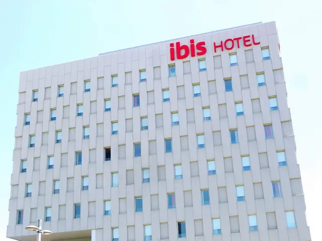 The image shows the ibis hotel in Berlin, Germany. It is a large building with windows, a street...