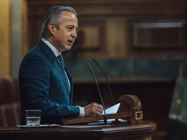 The image shows a man in a suit and tie standing at a podium in front of a microphone, with a glass...