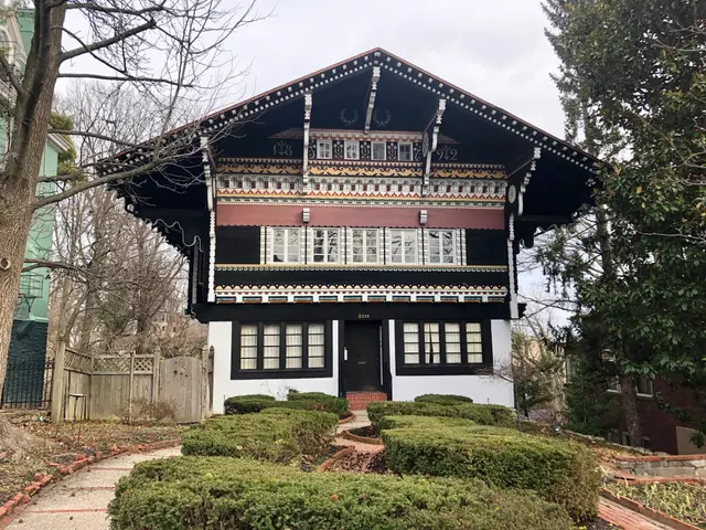 The image shows a black and white house with a tree in front of it, surrounded by plants, a...