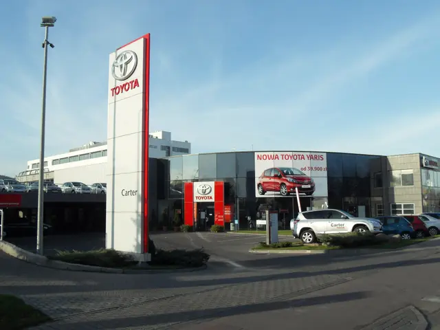 The image shows a Toyota dealership with cars parked in front of it, surrounded by buildings,...