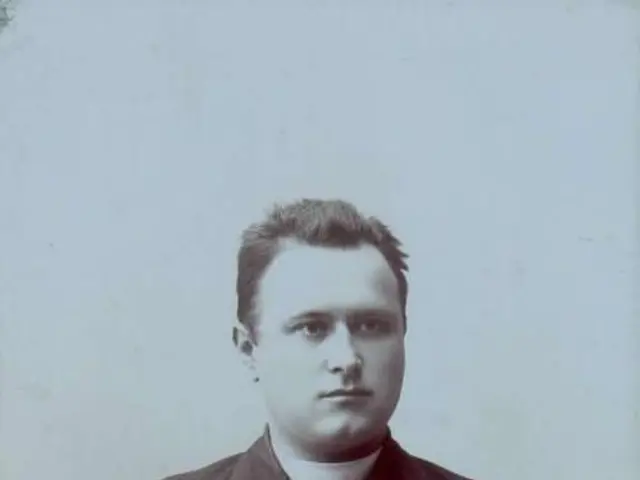 The image shows an old black and white photo of a man in a priest's robes, believed to be the first...