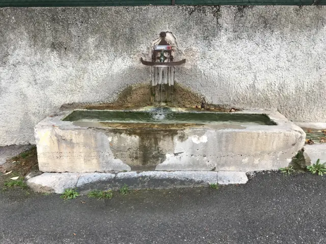 The image shows a small water fountain sitting on the side of a road, with a wall in the background...