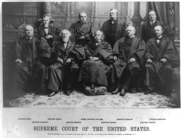 The image shows a black and white photo of a group of men sitting and standing in a room with a...