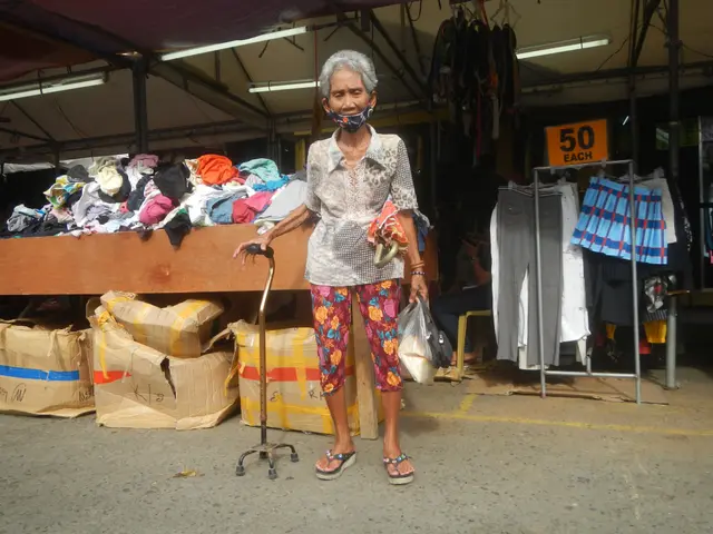 The image shows an elderly woman wearing a face mask standing in front of a market stall. She is...