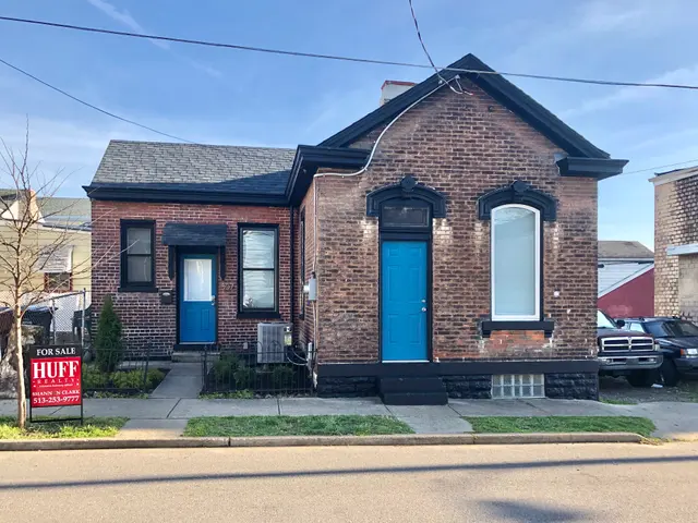The image shows a brick house with a blue door on the corner of a street, surrounded by buildings,...