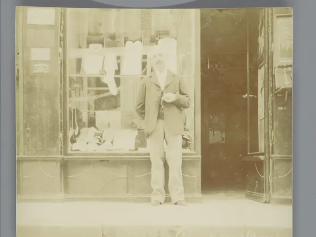 The image shows an old black and white photo of a man standing in front of a store. He is wearing a...