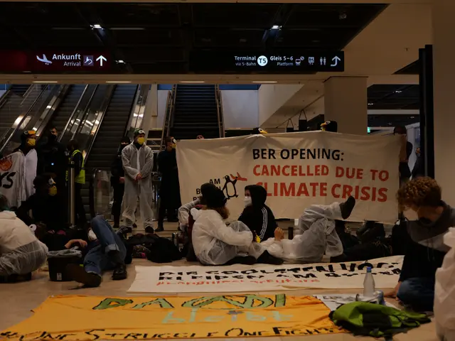 The image shows a group of people sitting on the floor of an airport, holding a banner that reads...