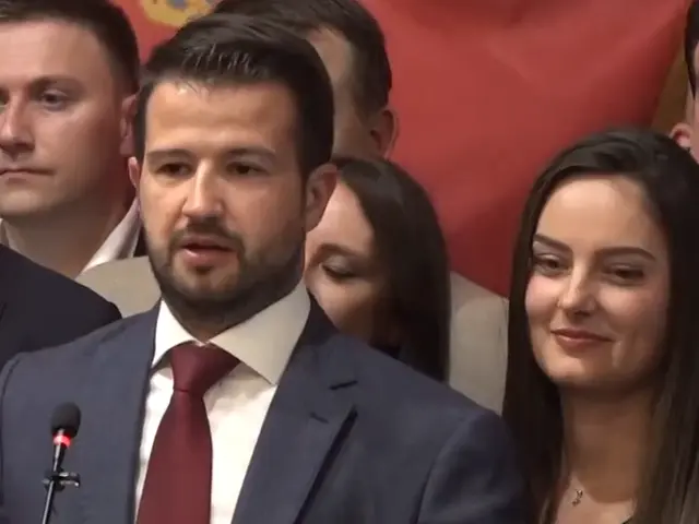 The image shows a man in a suit and tie standing at a podium in front of a crowd, with a microphone...