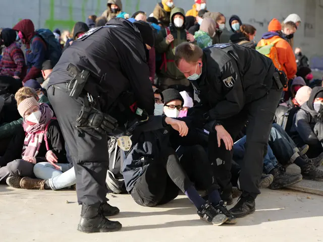 The image shows a group of people sitting on the ground next to each other, some wearing caps and...