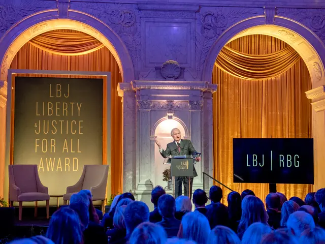 The image shows a man standing at a podium in front of a crowd of people sitting in chairs. On the...