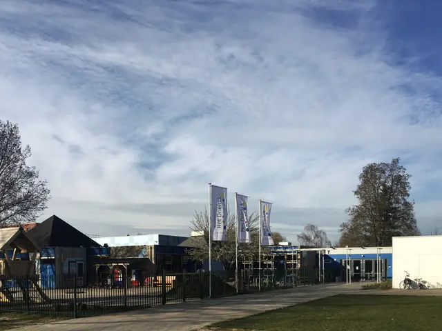 The image shows a large building with a lot of grass in front of it, surrounded by a metal fence, a...