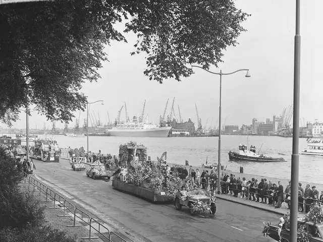 The image shows a black and white photo of a parade on a city street, with vehicles on the road,...