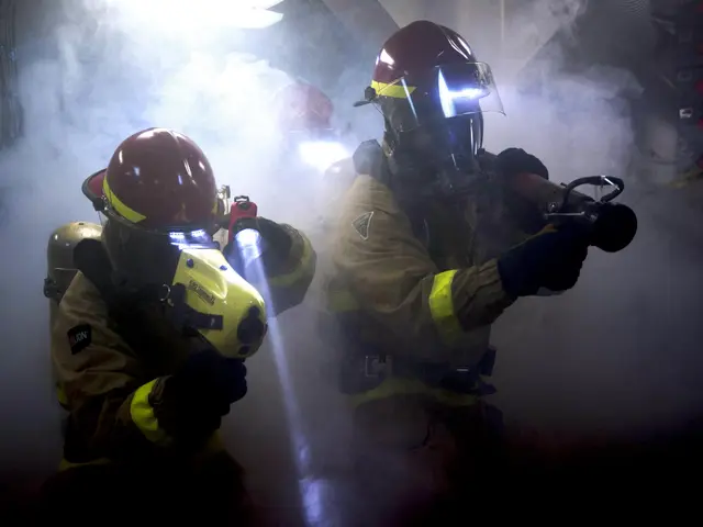 The image shows two firefighters in protective gear, wearing helmets and gas masks, spraying water...