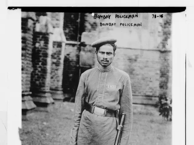 The image shows an old black and white photo of a bombay policeman standing on the ground, holding...