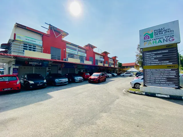 The image shows a parking lot with cars parked in front of a building, surrounded by trees and a...