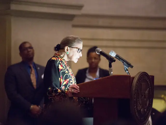 The image shows a woman standing at a podium in front of a crowd, with two microphones on the...
