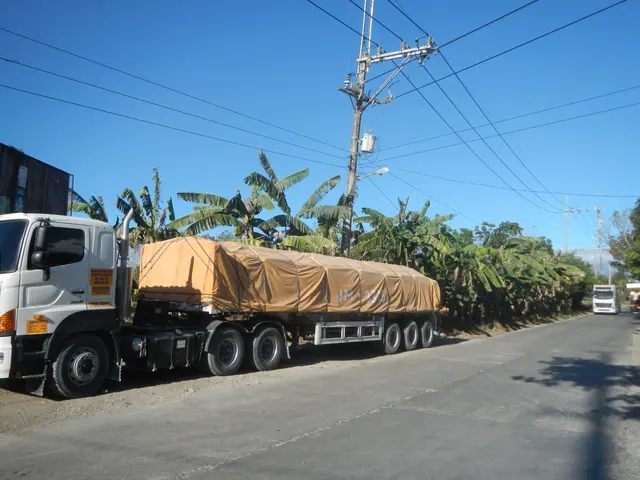 The image shows a truck with a tarp on the back of it driving down a road surrounded by trees,...