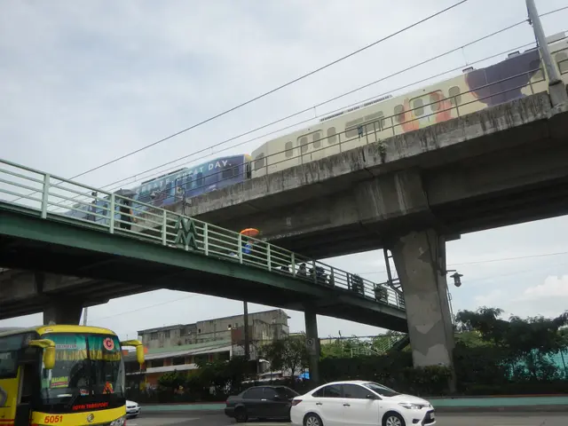 The image shows a yellow bus driving down a street next to a train on a bridge. The bridge has...