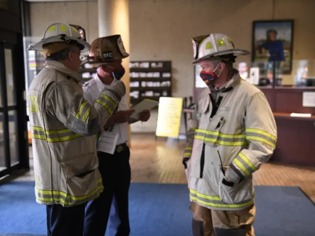 The image shows three firefighters wearing helmets and masks talking to each other in a room with a...