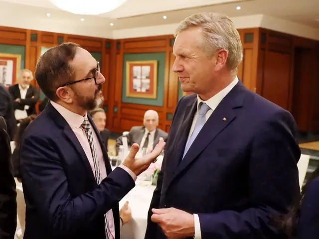 The image shows two men in suits talking to each other in a room with a group of people standing on...