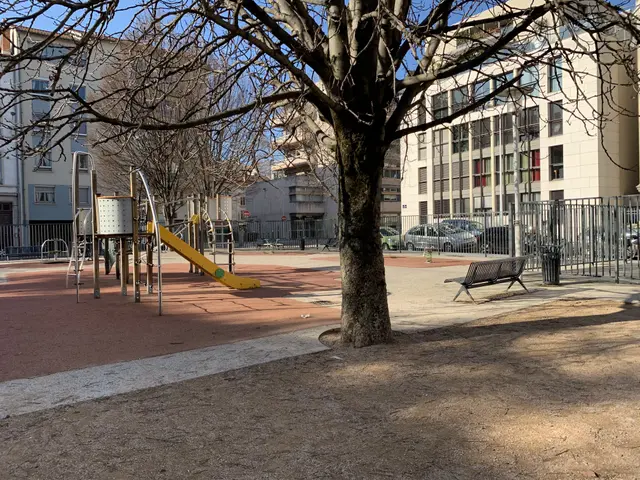 The image shows a playground with a yellow slide in the middle of it, surrounded by a metal fence,...