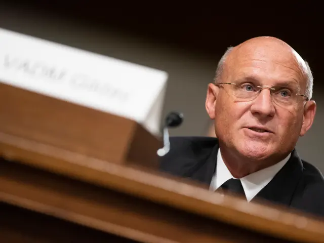 The image shows a man in a suit and tie sitting at a table with a name board and microphone in...