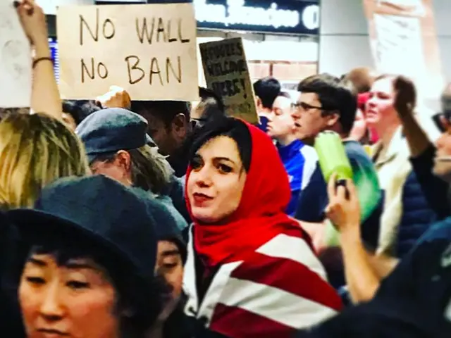 The image shows a group of people standing in front of a building, holding signs that read "No Wall...