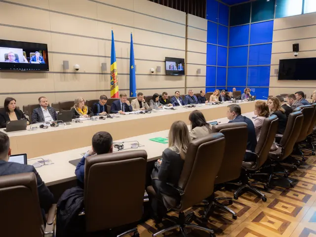 The image shows a large group of people sitting around a long table with laptops, mics, papers, and...