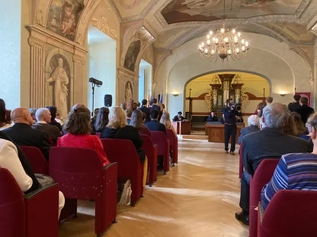 The image shows a group of people sitting in red chairs in a room with a wall adorned with photo...