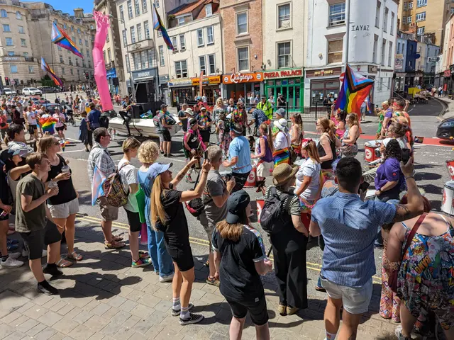 The image shows a crowd of people standing on the side of a street, some of them holding flags,...