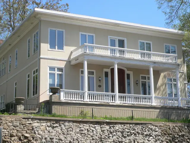 The image shows a large white house with a stone wall in front of it, windows, doors, pillars,...
