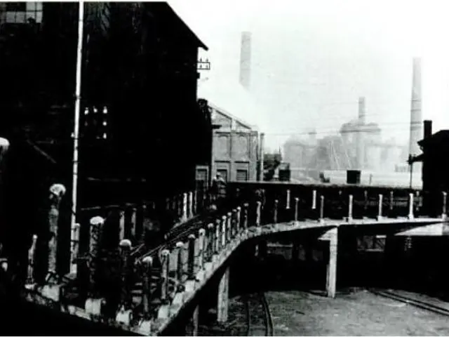 The image shows a black and white photo of a bridge over a train track, with buildings on either...