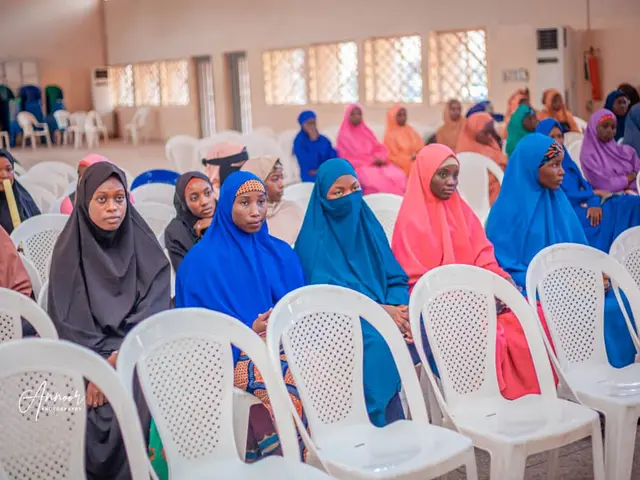 The image shows a group of women sitting in chairs in a room with a wall in the background, windows...