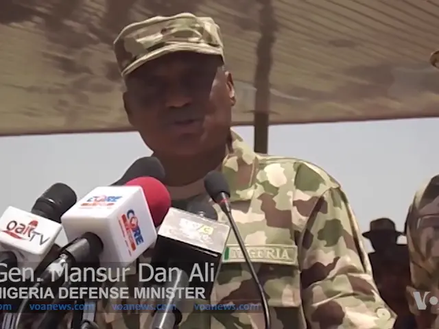 The image shows a man in military uniform standing at a podium with microphones in front of him,...