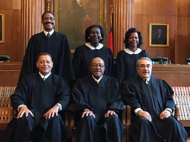 The image shows a group of people sitting in chairs in a courtroom, wearing black robes and...