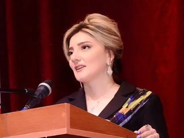 The image shows a woman standing at a podium in front of a microphone, with a red curtain in the...