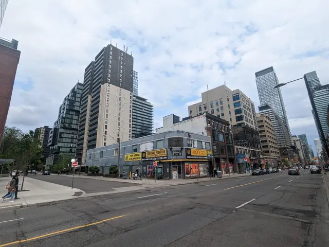 The image shows a city street with tall buildings in the background, vehicles on the road, people...