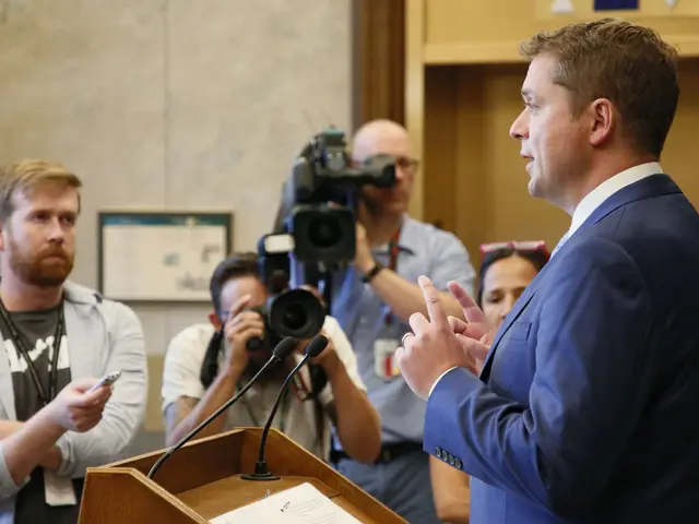 The image shows a man in a suit and tie standing at a podium in front of a group of people, some of...