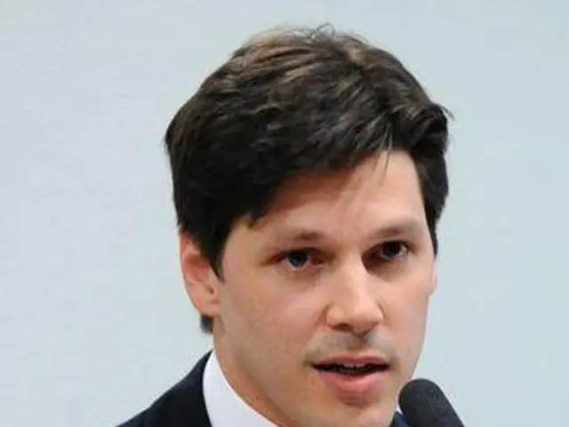 The image shows a man in a suit and tie speaking into a microphone in front of a wall. He appears...