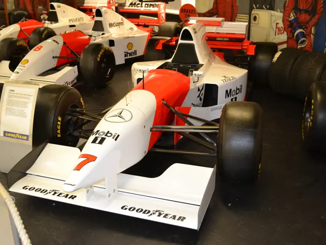 The image shows a blue race car on display in a museum, with a red background. The car is on a...