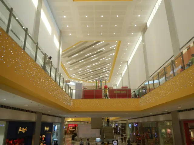 The image shows the interior of a shopping mall with people walking around, stores with name...