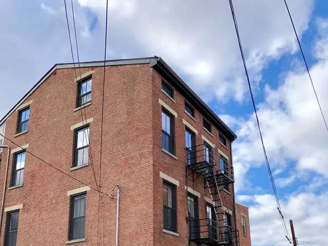 The image shows a brick building with windows, steps with railings, and cars parked in front of it....