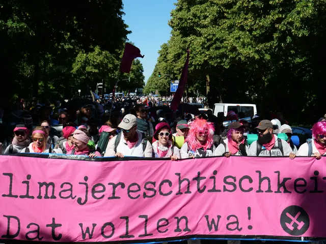 The image shows a group of people walking down a street, holding a pink banner with the words...