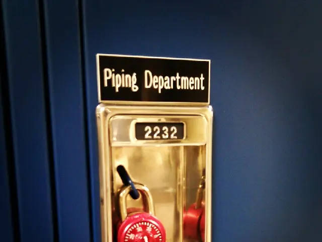 The image shows a blue locker with a red padlock on it and a name board with the words "Piping...