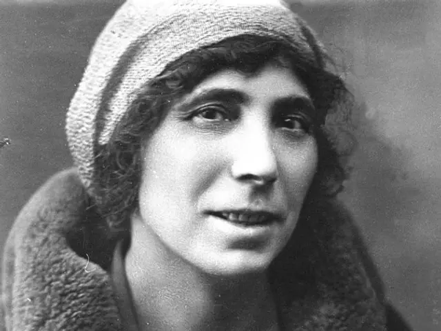 The image shows a black and white photo of a woman wearing a fur coat and a cap, with a blurred...