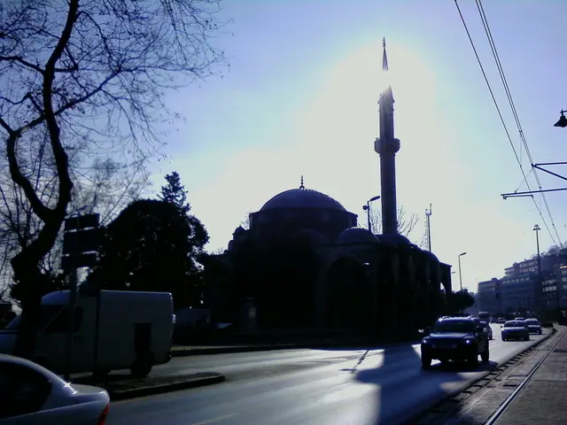 The image shows a mosque in the middle of a city street, surrounded by buildings, street poles,...